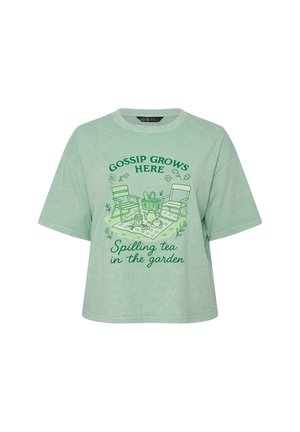 T-shirt corta verde chiaro con illustrazione verde di due sedie, cesta da picnic, servizio da tè e testo "Il pettegolezzo cresce qui, versando il tè in giardino."