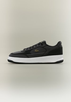 Baskets basses noires avec semelle blanche, logo crocodile doré de Lacoste sur le côté, lacets noirs et talon texturé, posées sur un fond neutre.