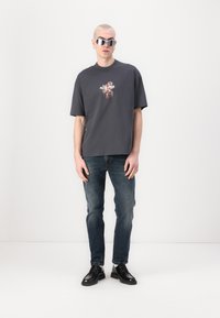 Camiseta gris de gran tamaño con diseño floral bordado, combinada con jeans azules y zapatos negros con cordones. El modelo lleva unas gafas de sol blancas.