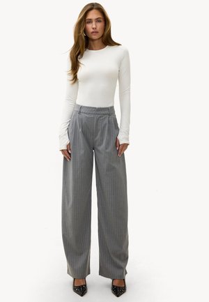 Femme debout portant un haut blanc à manches longues, un pantalon ample à rayures grises taille haute, et des chaussures pointues noires cloutées.