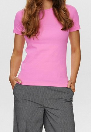 Femme aux longs cheveux bruns portant une chemise rose à manches courtes et un pantalon gris à fines rayures, les mains dans les poches.