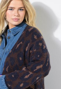 Gilet en imprimé léopard marron et bleu marine avec une texture douce, porté par-dessus une chemise en denim bleu. Caractéristiques : épaules tombantes et coupe ample.
