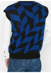 Gilet in maglia senza maniche con audace motivo geometrico a zigzag blu e nero, indossato sopra una camicia bianca a maniche lunghe e jeans blu.