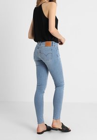 Vaqueros de mezclilla delgada en un azul claro con un parche de cuero en la parte posterior de la cintura, combinados con una blusa sin mangas negra y sandalias negras tipo deslizar.