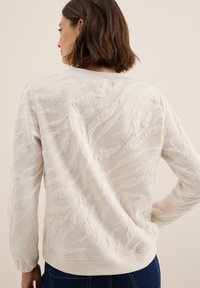 Sweat-shirt couleur crème avec des motifs texturés et ondulés, des poignets et un col côtelés, offrant une coupe décontractée et un tissu doux et tissé.