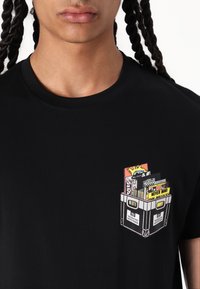 Black cotton t-shirt featuring a printed graphic of a crate with colorful book covers in the chest pocket area.