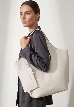 Vrouw met een donker colbert die een grote witte leren totebag draagt met een aangehechte witte ritstasje aan haar schouder tegen een effen achtergrond.
