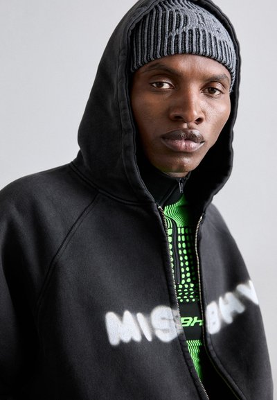 Schwarze Zip-up-Hoodie mit einer engen Kapuze, auf der ein weißer "MISBHV"-Aufdruck zu sehen ist. Darunter ist ein grünes, strukturiertes Shirt mit einem Punktmuster sichtbar.