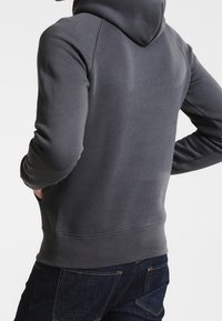Felpa con cappuccio di colore grigio scuro, caratterizzata da una texture morbida, tasca a marsupio e polsini a costine. I dettagli delle cuciture visibili arricchiscono il design.