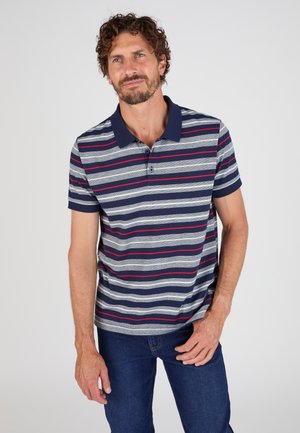 Uomo con barba che indossa una polo a righe blu navy, grigie e rosse e jeans blu, in piedi davanti a uno sfondo chiaro e uniforme.