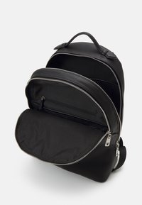 Un sac à dos en cuir noir avec une texture lisse, doté d'une grande ouverture zippée, d'une petite poche avant et d'accents en métal argenté.