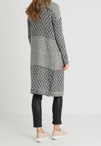 Lång grå och svart mönstrad cardigan med ett geometriskt mönster, kombinerad med svarta leggings och ljusrosa sneakers. Slen yta.