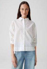 Witte lange mouwen shirt met knoopsluiting, voorzien van geborduurde details op de mouwen, gecombineerd met lichtblauwe jeans. Glad materiaal, ontspannen pasvorm.