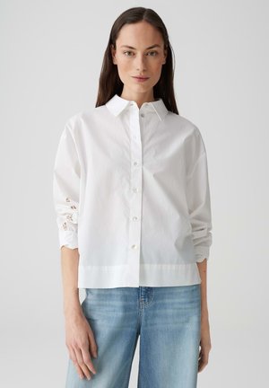 Witte lange mouwen shirt met knoopsluiting, voorzien van geborduurde details op de mouwen, gecombineerd met lichtblauwe jeans. Glad materiaal, ontspannen pasvorm.