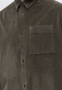 Chemise en velours côtelé marron avec des boutons sur le devant et une poche poitrine carrée sur le côté gauche.