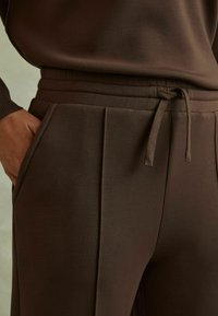 Bruine sweatpants met een gladde textuur, voorzien van een koordin de taille, zijzakken en subtiele plooien aan de voorkant.