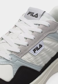 Κοντινό πλάνο λευκού, γκρι και μαύρου αθλητικού παπουτσιού FILA με λευκά κορδόνια και πάνελ από σουέντ, εμφανίζοντας το λογότυπο της μάρκας στη γλώσσα και στο πλάι.