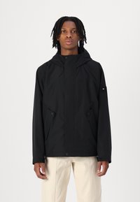 Schwarze Kapuzenjacke mit einem schlanken Design, weicher Textur, Druckknopfverschlüssen und seitlichen Taschen; dezentes Branding auf dem Ärmel.