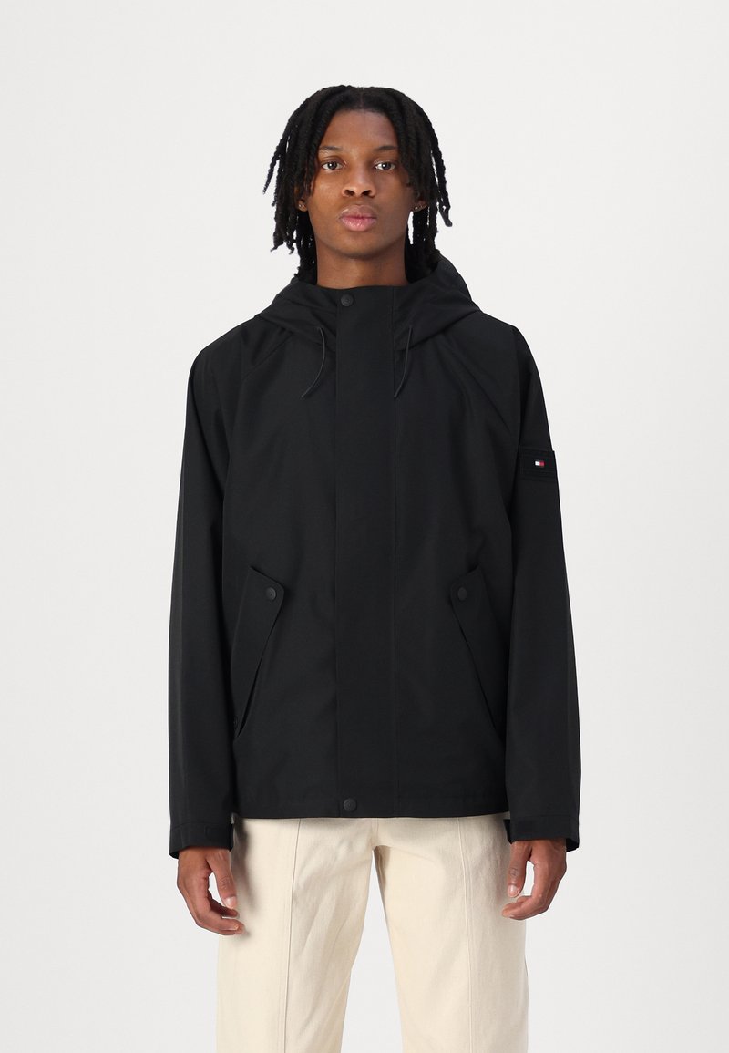 Schwarze Kapuzenjacke mit einem schlanken Design, weicher Textur, Druckknopfverschlüssen und seitlichen Taschen; dezentes Branding auf dem Ärmel.