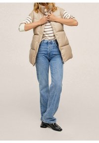 Beige padded vest with a high collar, worn over a striped shirt. Paired with light blue high-waisted jeans and black shoes.