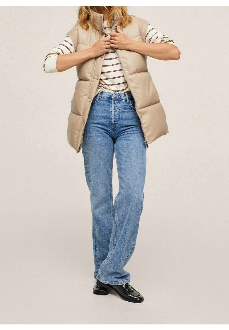Beige padded vest with a high collar, worn over a striped shirt. Paired with light blue high-waisted jeans and black shoes.
