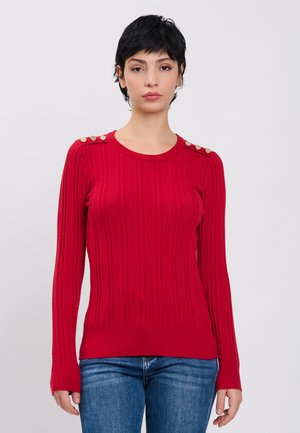 Junge Frau mit kurzen schwarzen Haaren, die einen roten Rippstrickpullover mit goldenen Knöpfen auf den Schultern und blaue Jeans vor schlichtem Hintergrund trägt.