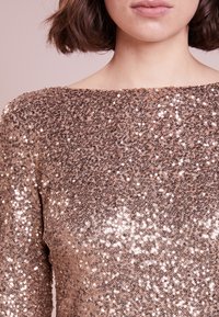 Kvinna med kort brunt hår iklädd en långärmad topp med rosaguldiga paljetter och bred ringning mot en ljusrosa bakgrund.