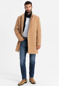 Abrigo de lana marrón claro, bufanda oscura, camisa de rayas claras, jeans azules y botas de tobillo marrón claro. El abrigo tiene un diseño recto con un acabado limpio.