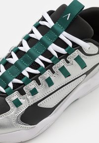 Sapatilha com um upper em malha preta, detalhes em pele prateada, atacadores texturizados verdes e atacadores sintéticos brancos; apresenta um design de onda único.