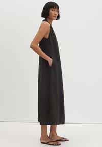 Femme debout portant une robe midi noire sans manches à col halter avec poches, chaussée de sandales noires type tongs sur un fond uni.