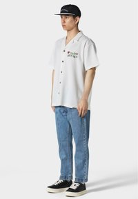 Camicia bianca a maniche corte con bottoni e ricami colorati sul petto, abbinata a jeans azzurri e sneaker nere. Un cappellino nero completa il look.