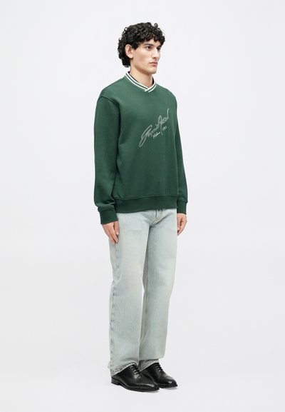 Πράσινο φούτερ με λευκό κεντητό σχέδιο, ριμπ γιακά και μακριά μανίκια. Συνδυασμένο με τζιν ανοιχτού τόνου και μαύρα παπούτσια.