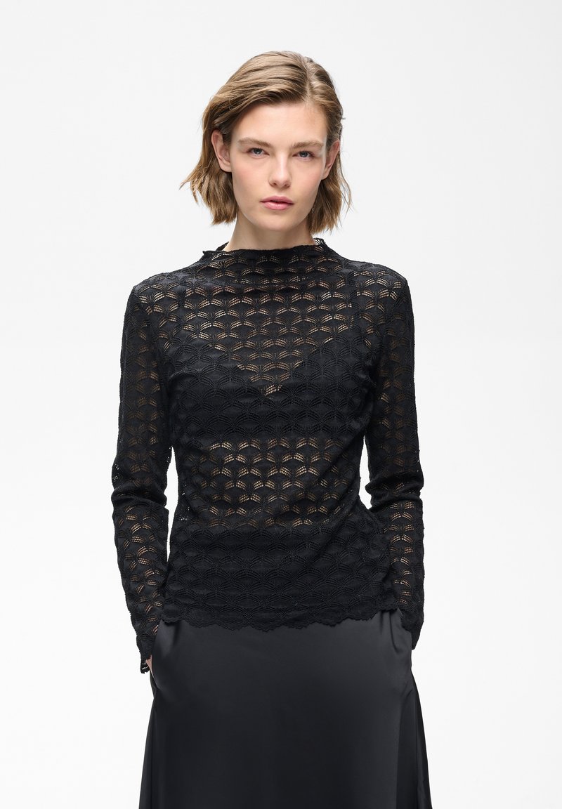 Femme aux cheveux courts portant un top en dentelle noire à manches longues par-dessus un caraco noir, assorti à une jupe en satin noire, les mains dans les poches.