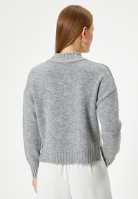 Grauer Strickpullover mit rippenförmigem Kragen und Saum, oversized Schnitt, tiefer gelegte Schultern und strukturierte Oberfläche. Die Rückansicht zeigt ein hervorgehobenes Strickmuster.