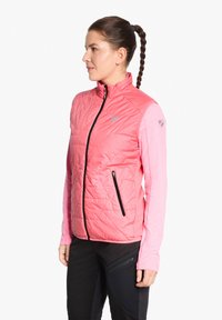 Rosa gepolsterte Weste mit hohem Kragen, zwei schwarzen Reißverschluss-Taschen und einem Logo. Lange rosa Ärmel sind darunter sichtbar, kombiniert mit schwarzen Hosen.