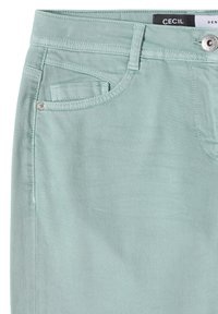 Jean en denim vert clair en gros plan montrant la poche avant, la poche à pièces, la boucle de ceinture et le bouton en métal avec l'étiquette de la marque à l'intérieur de la taille.