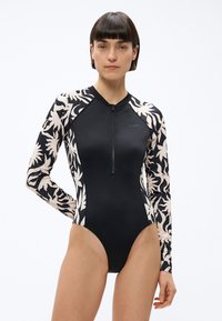 Žena oblečená v čiernych jednodielnych plavkách s dlhým rukávom, s béžovými rukávmi s kvetinovým vzorom a predným zipsom, stojaca na bielom pozadí.