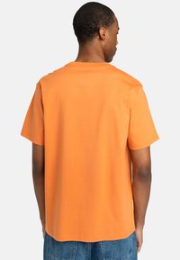 T-shirt orange à manches courtes en coton doux, présentant une coupe décontractée et un col rond. Détails de couture le long des épaules.