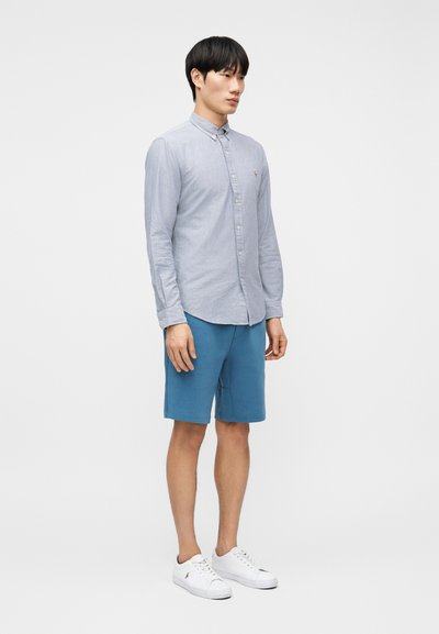 Άνδρας που στέκεται με γαλάζιο πουκάμισο με κουμπιά, βερμούδα μέντας μέχρι το γόνατο και λευκά αθλητικά παπούτσια σε απλό λευκό φόντο.