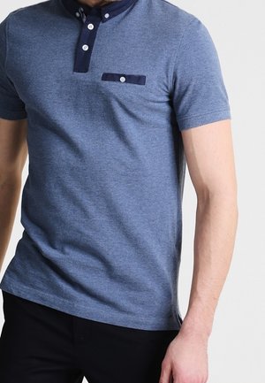 Mann trägt ein blaues kurzärmeliges Poloshirt mit dunklem Besatz und Knöpfen, kombiniert mit schwarzen Hosen, steht vor einem einfarbigen Hintergrund.