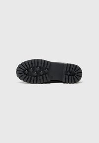 Semelle en caoutchouc noir avec des rainures profondes, des motifs circulaires et rectangulaires, dotée d'un talon plat et d'une zone de renforcement au niveau des orteils pour plus de durabilité.