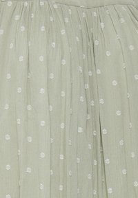 Un tissu vert clair avec une texture diaphane présente des pois brodés blancs et de doux plis, créant une apparence douce et fluide.