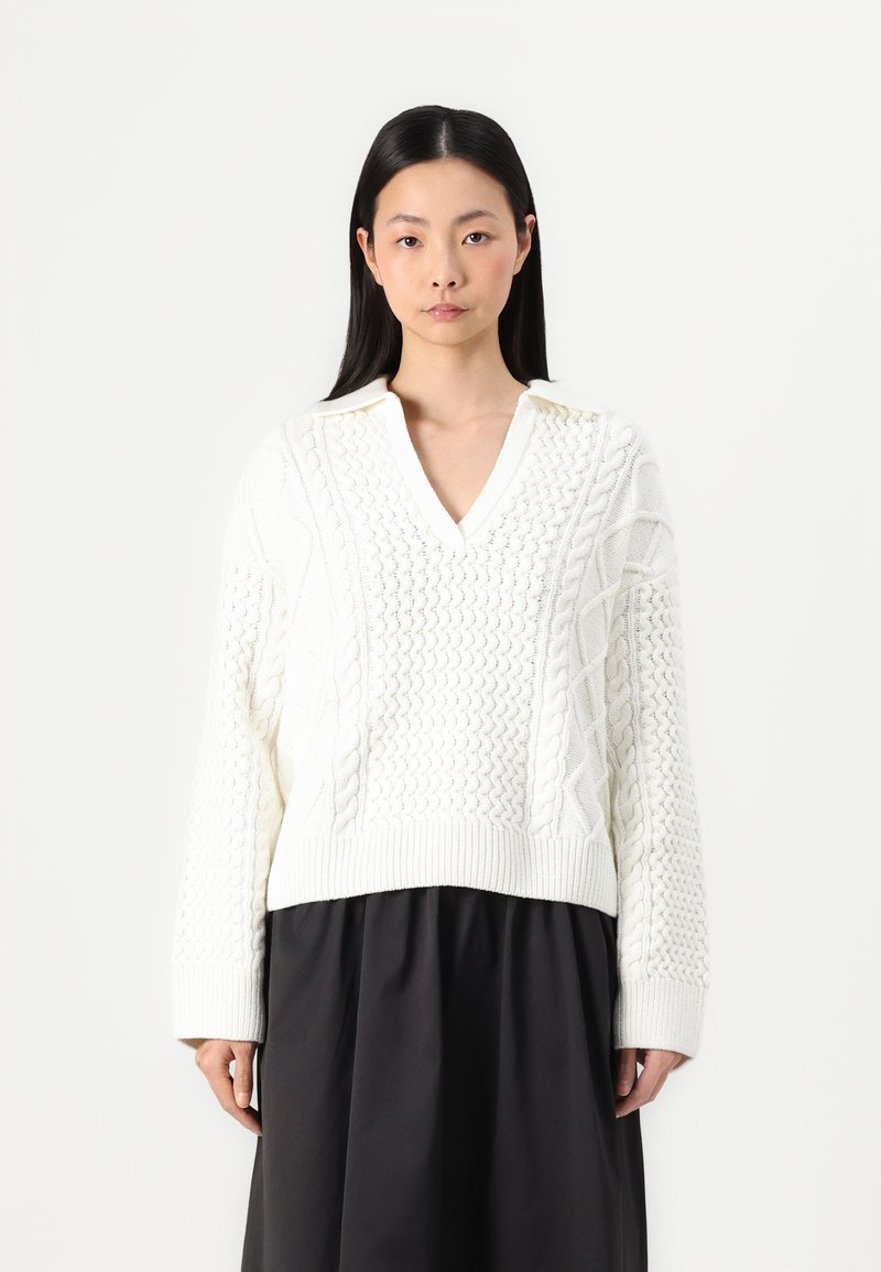 Hvid kabelstrikket sweater med V-udskæring og lange ærmer. Har teksturerede mønstre og ribbet kant. Bæres over en mørk nederdel.