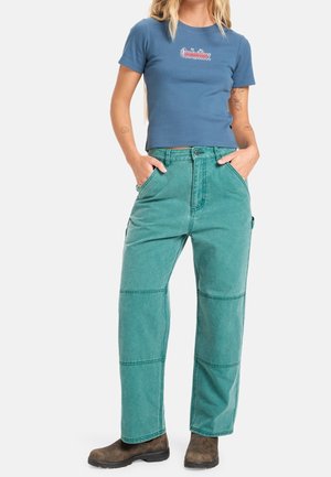 Kobieta ubrana w dopasowany niebieski t-shirt i wysokie spodnie koloru teal z brązowymi butami, ręce w kieszeniach, stoi na białym tle.