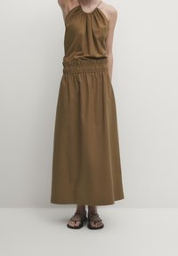 Robe longue marron à encolure licou avec un corsage froncé et une taille élastique. Le modèle l'a associée à des sandales noires à fines brides.