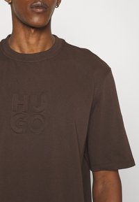T-shirt en coton marron à manches courtes, col rond, avec un logo "HU GO" en relief dans une teinte légèrement plus foncée sur le devant.