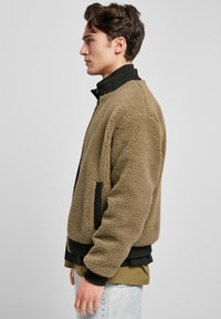 Kosmata jakna olivno zelene barve s črnimi poudarki, rebrastim ovratnikom in stranskimi žepi. Mehka tekstura z ohlapnim, presežnim krojem.