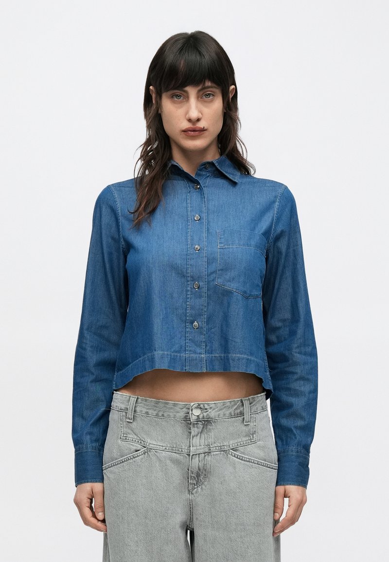 Chemise en denim coupée en bleu moyen avec un col boutonné, une poche poitrine et des manches longues. Associée à un jean gris taille haute.