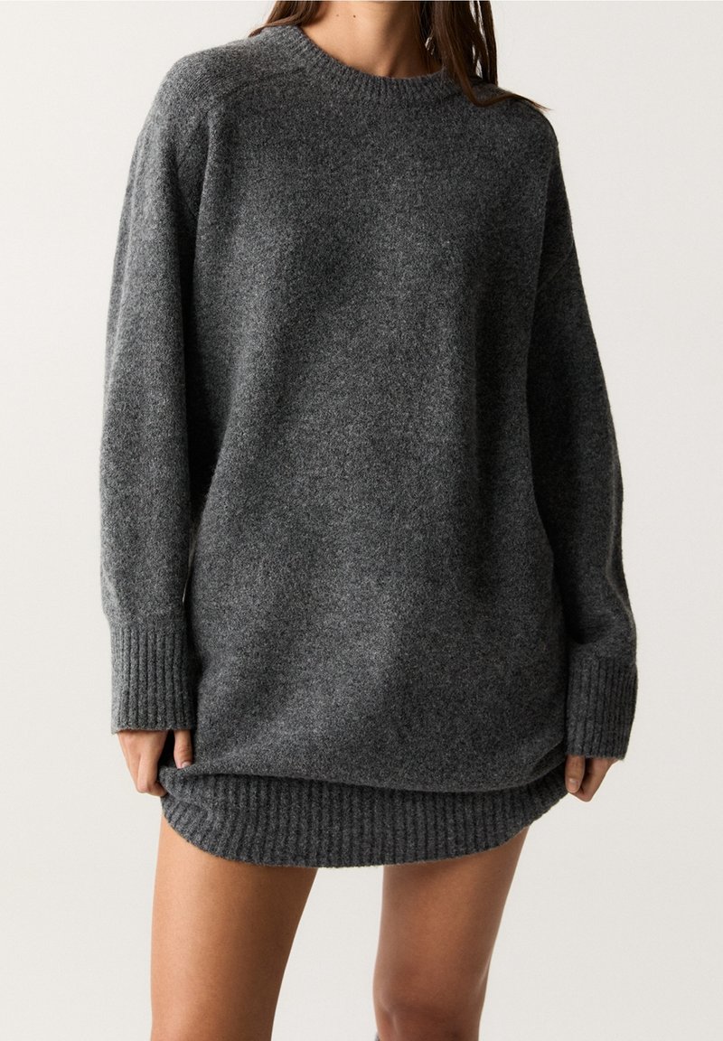 Femme portant une robe-pull tricotée oversize gris foncé avec poignets et ourlet côtelés, debout devant un fond clair uni.