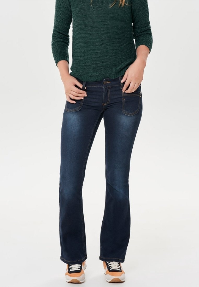 Jean flare bleu foncé en denim, avec des poches avant, une passante de ceinture et des coutures contrastées. Associé à un pull vert et des baskets.